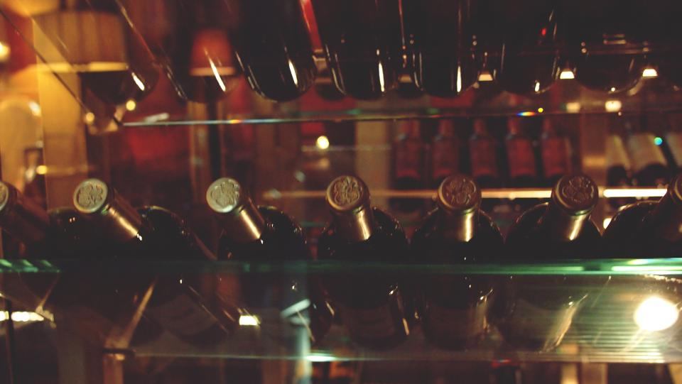 Rows of dark wine bottles with wax capsules resting on glass cellar shelves.